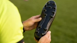 Mãos segurando o solado de uma chuteira com travas moldadas em campo de grama