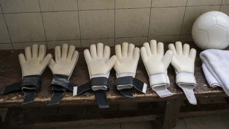 Três pares de luvas de goleiro lado a lado num banco de madeira, palmas para cima com texturas de látex diferentes